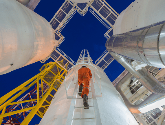 A worker in an orange jumpsuit climbs a ladder on a white industrial structure against a dark blue sky.